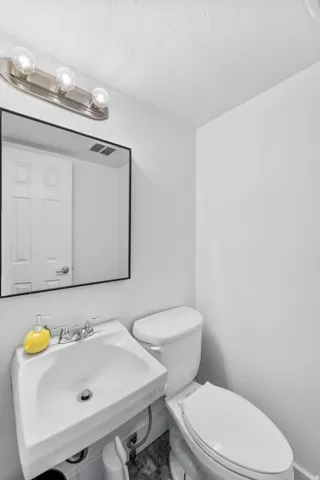 Pedestal sink, wall-mounted mirror, brushed nickel light fixture, toilet, and dark-toned flooring