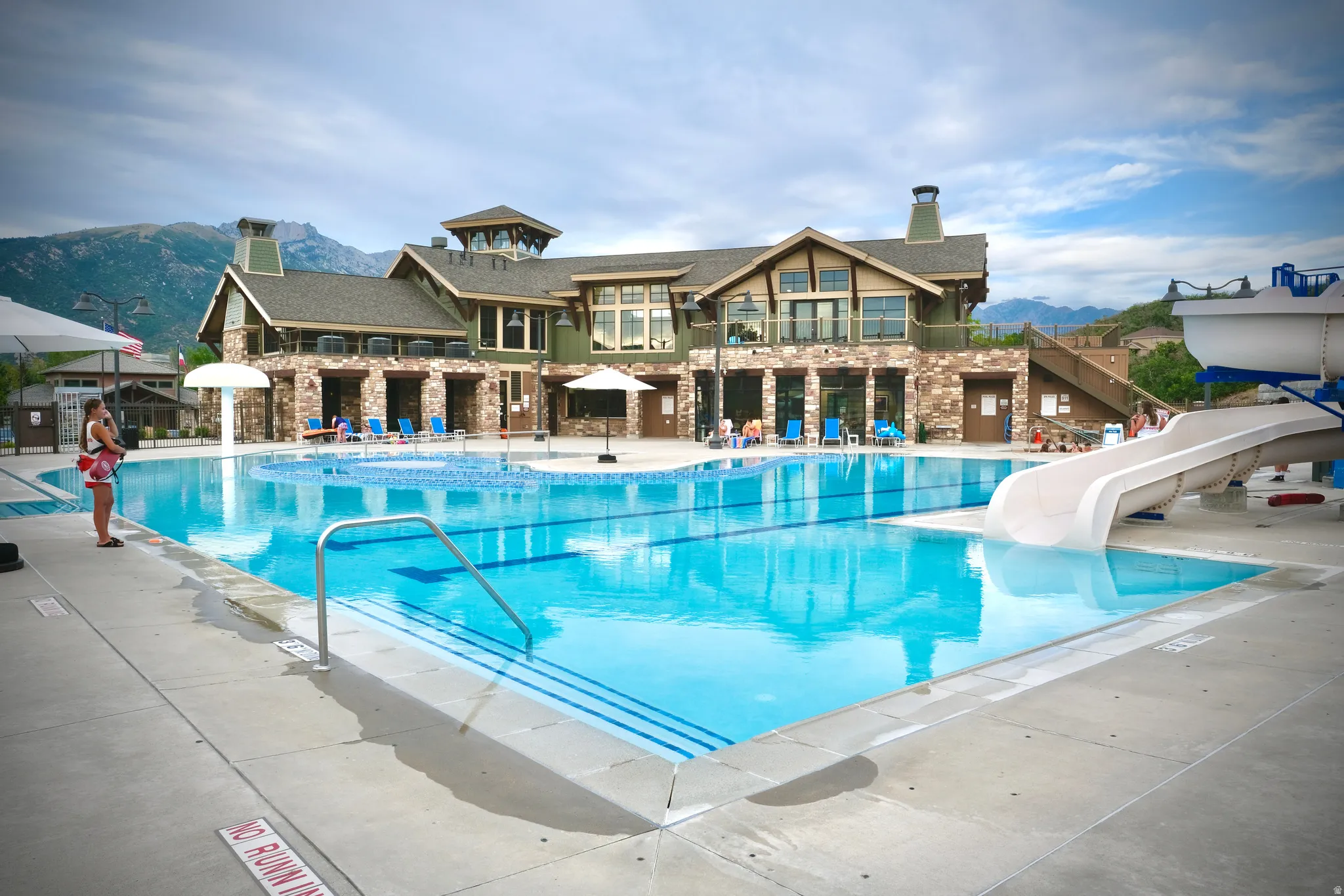Expansive outdoor swimming pool with a water slide and a separate pool area, complemented by a multi-level clubhouse featuring stone and wood-paneled exterior finishes, an upper-level balcony, and large windows