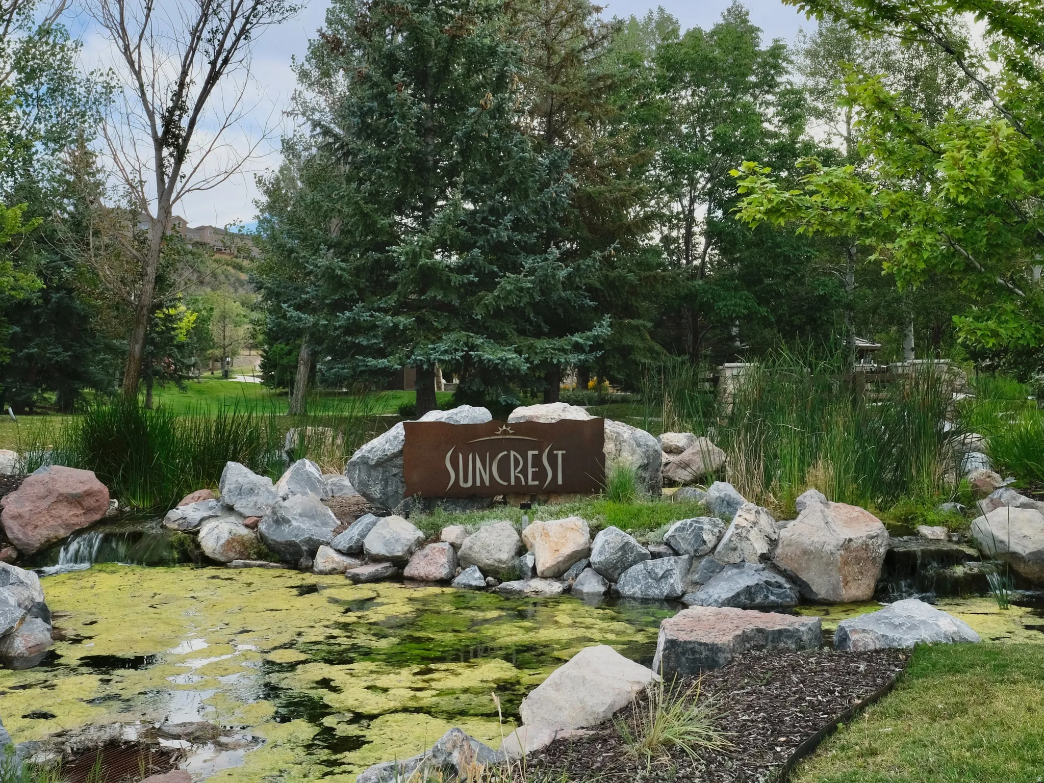 SunCrest clubhouse grounds, featuring a landscaped pond, mature trees, natural rock features, and the community’s signature SunCrest monument.