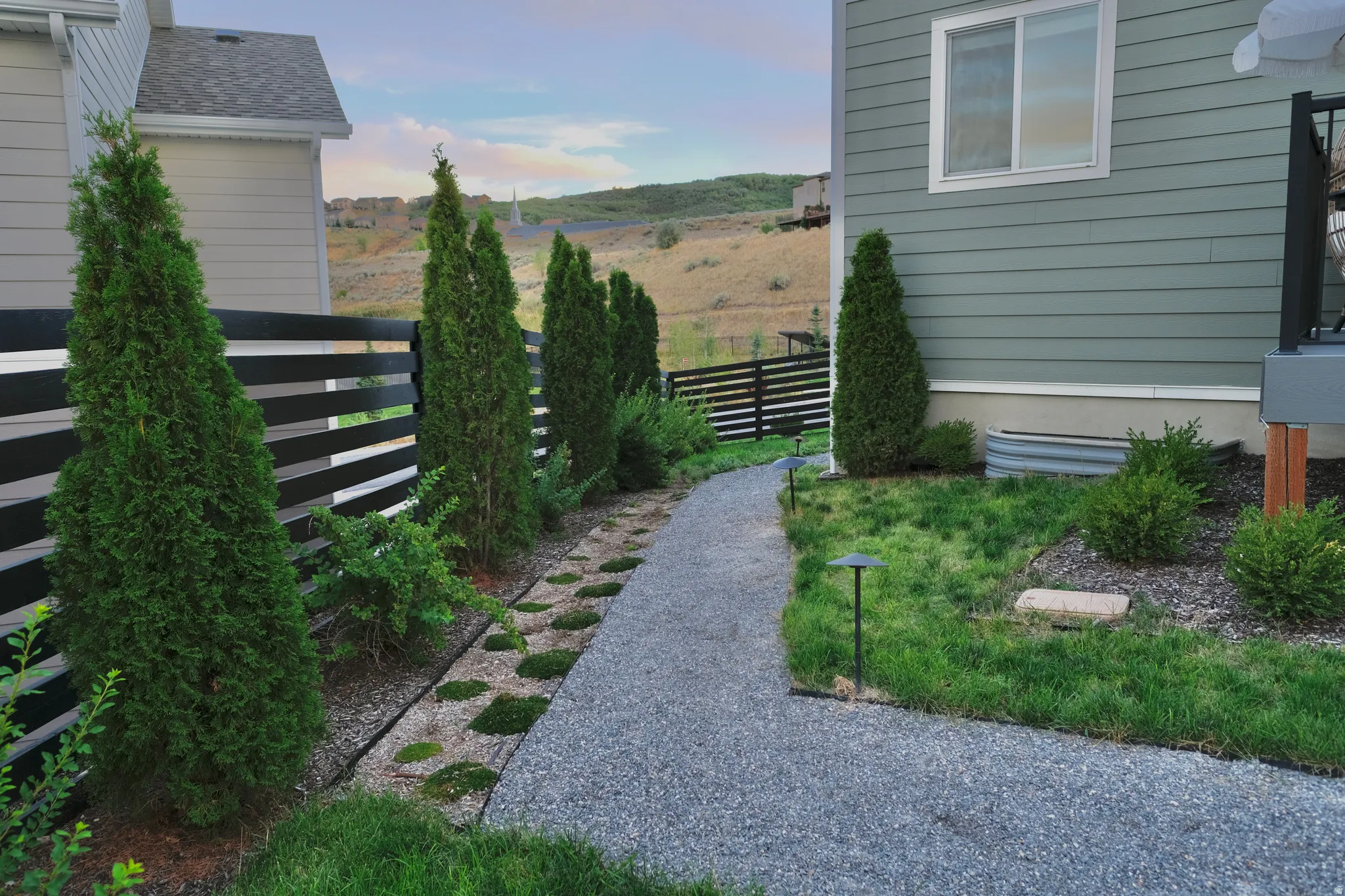 Gravel pathway bordered by landscaped beds, featuring a black horizontal slat fence and established evergreen trees