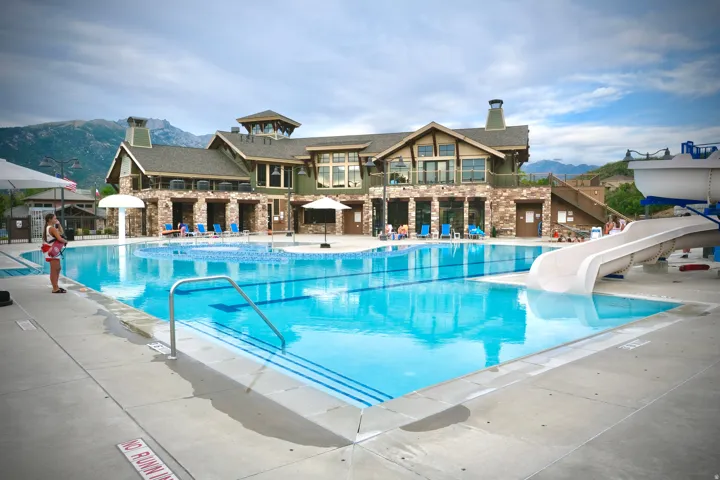 Expansive outdoor swimming pool with a water slide and a separate pool area, complemented by a multi-level clubhouse featuring stone and wood-paneled exterior finishes, an upper-level balcony, and large windows