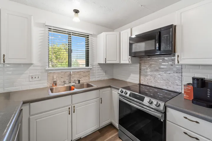 White cabinetry with bronze hardware, solid surface countertops, stainless steel double basin sink, white subway tile backsplash, and wood-finish flooring
