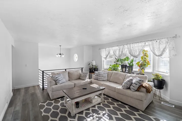Spacious living area featuring wood-finish flooring, a contemporary chandelier, large windows with blinds, and a metal horizontal bar railing