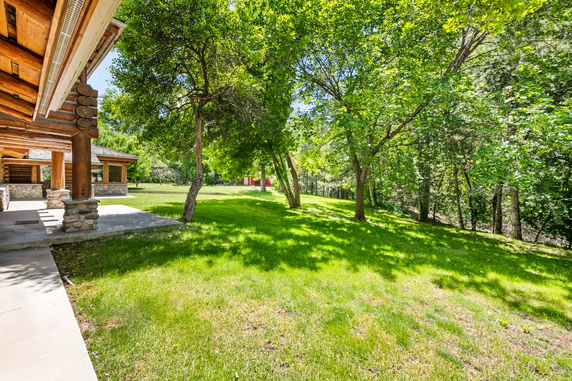 Expansive green lawn surrounded by mature trees, featuring a concrete patio with natural stone pillars and exposed log beams