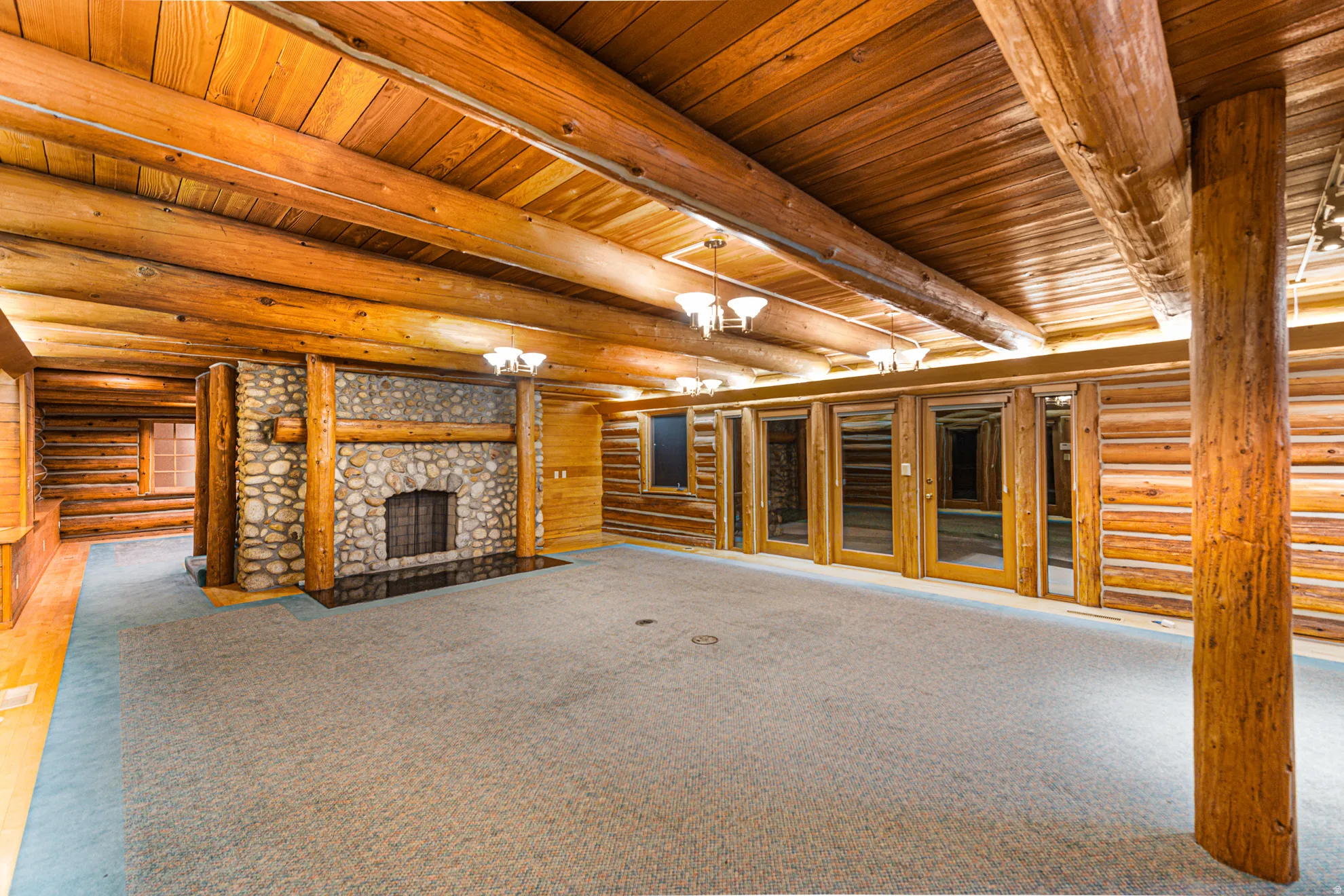 Log-paneled walls and ceiling beams, stone fireplace with a metal grate, carpeting, and multiple wood-framed glass doors