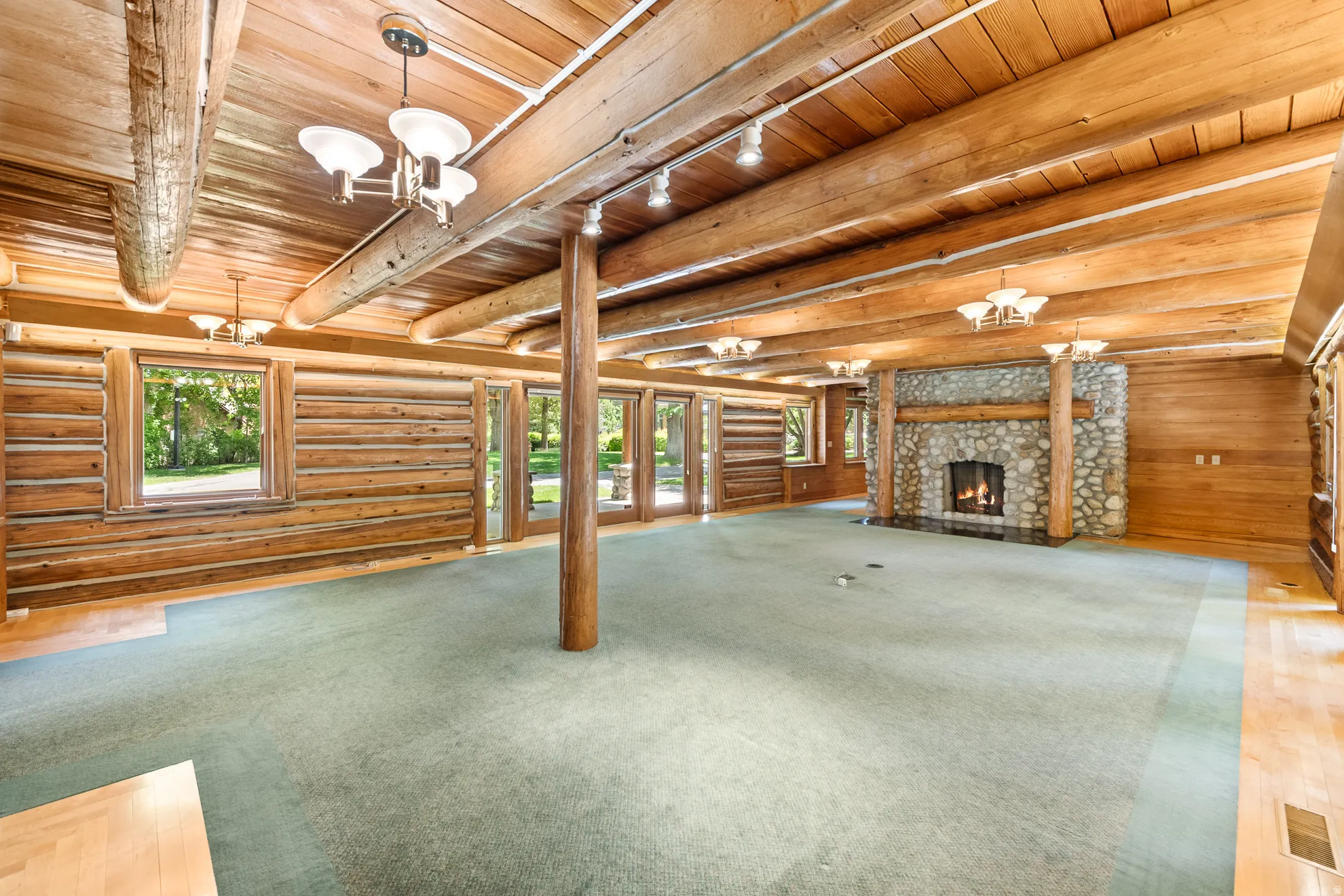 Spacious living area featuring exposed log beam ceiling, a stone fireplace with a substantial log mantel, light wood-finish flooring, and multiple windows with exterior views