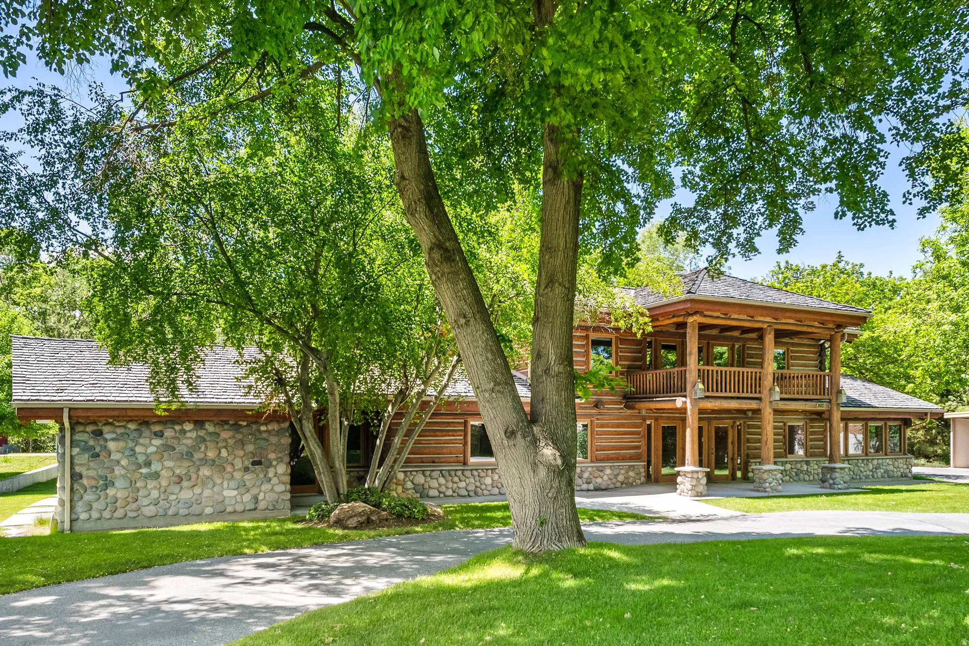Log cabin exterior featuring a multi-level facade with a stone foundation, a second-story balcony with wood railings, a covered entry porch, and expansive lawns