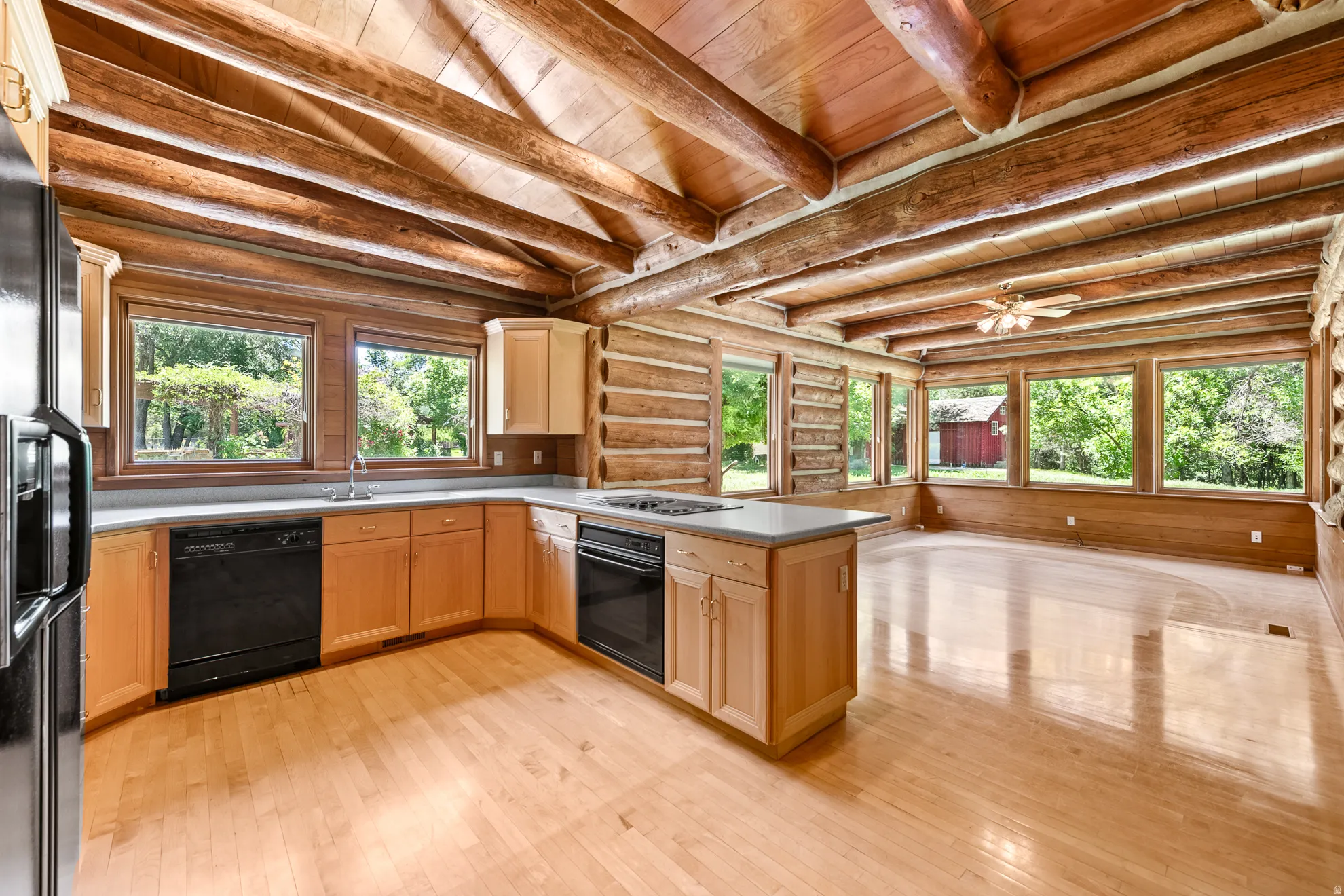 Open-concept kitchen and living area featuring exposed wood beams, light wood-finish flooring, natural wood cabinetry, and extensive window coverage