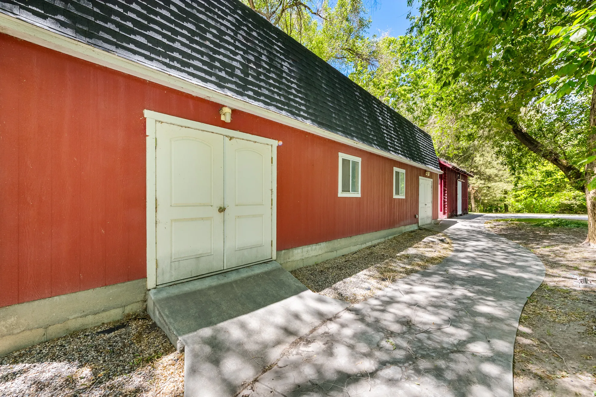 Red-sided structure featuring a dark shingle roof, white double doors with a concrete ramp, and multiple white-framed windows