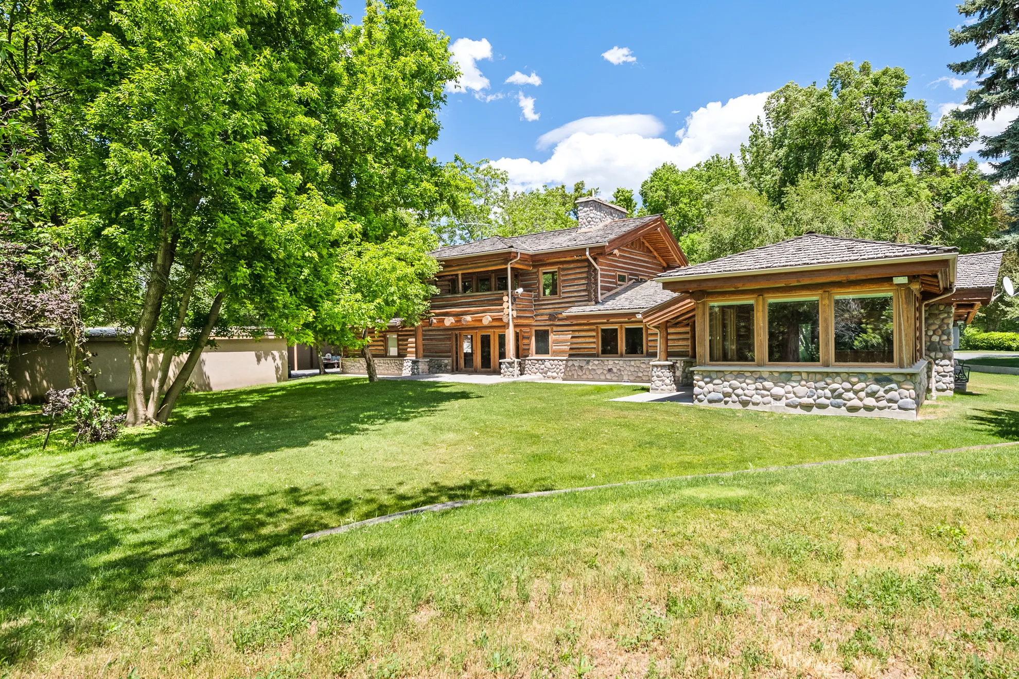 Expansive lawn surrounding a log home featuring a river rock foundation, multiple large windows, and a prominent wood-shingle roof