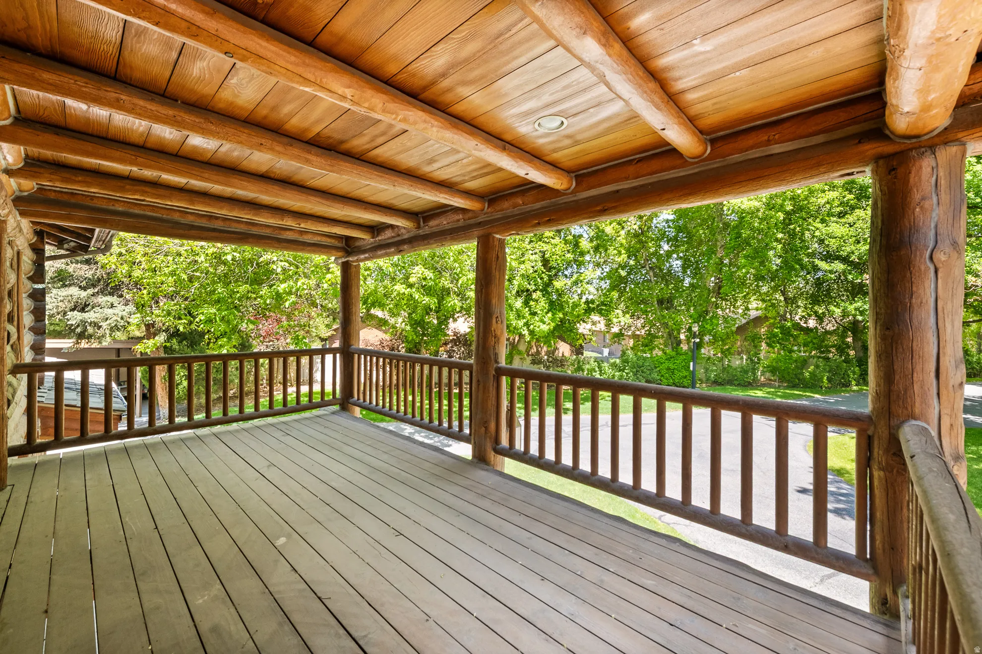Spacious covered porch featuring a natural wood ceiling, exposed log beams, wooden plank flooring, and a log railing