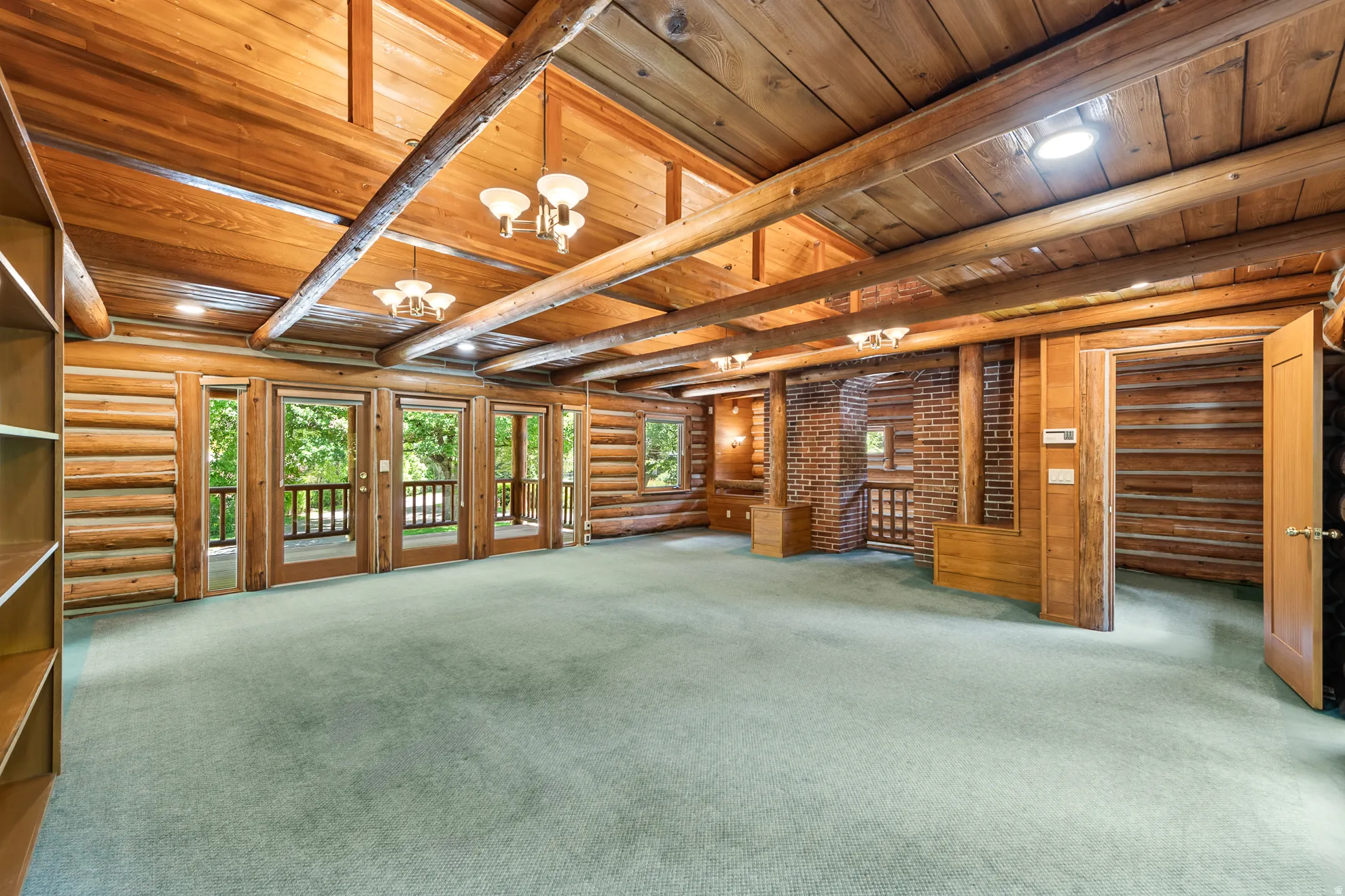 Spacious interior featuring exposed log walls, vaulted wood ceilings with exposed beams, recessed lighting, and multiple glass doors opening to an outdoor balcony