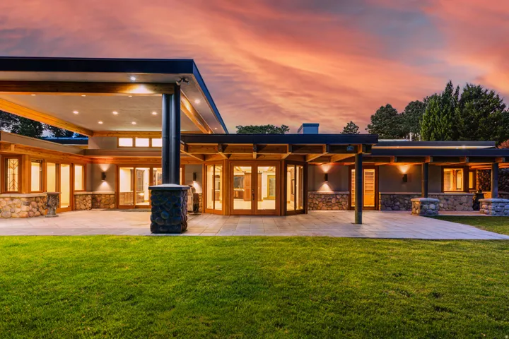 Expansive stone patio with a manicured lawn, multiple glass-paneled entryways, exposed wood beams, and recessed lighting