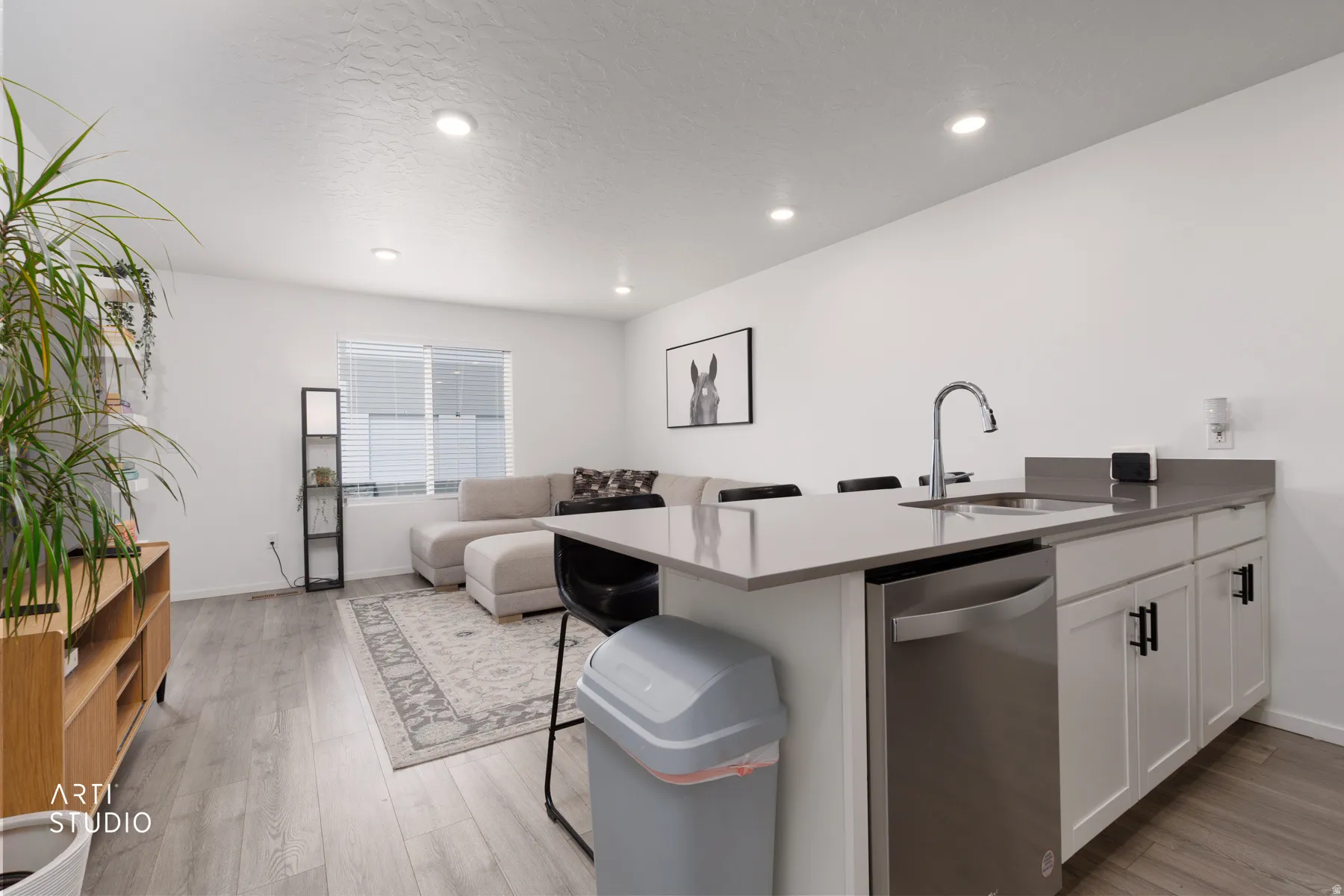 Open-concept living space featuring wood-finish flooring, recessed lighting, a kitchen island with an integrated sink, and white kitchen cabinetry with black hardware