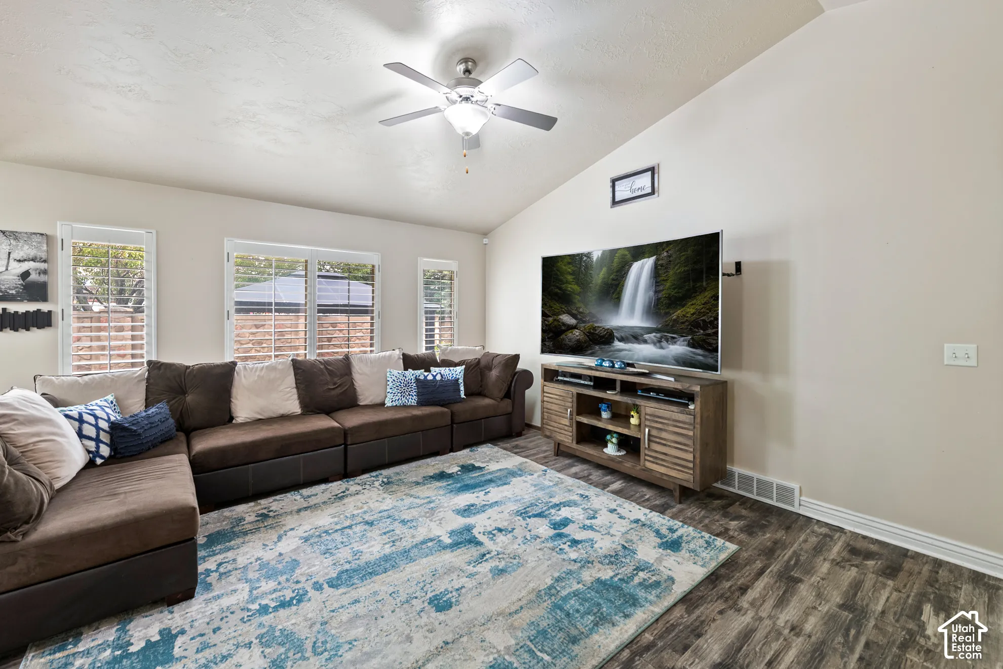 Vaulted ceiling with an attached ceiling fan, wood-finish flooring, and multiple windows with plantation shutters