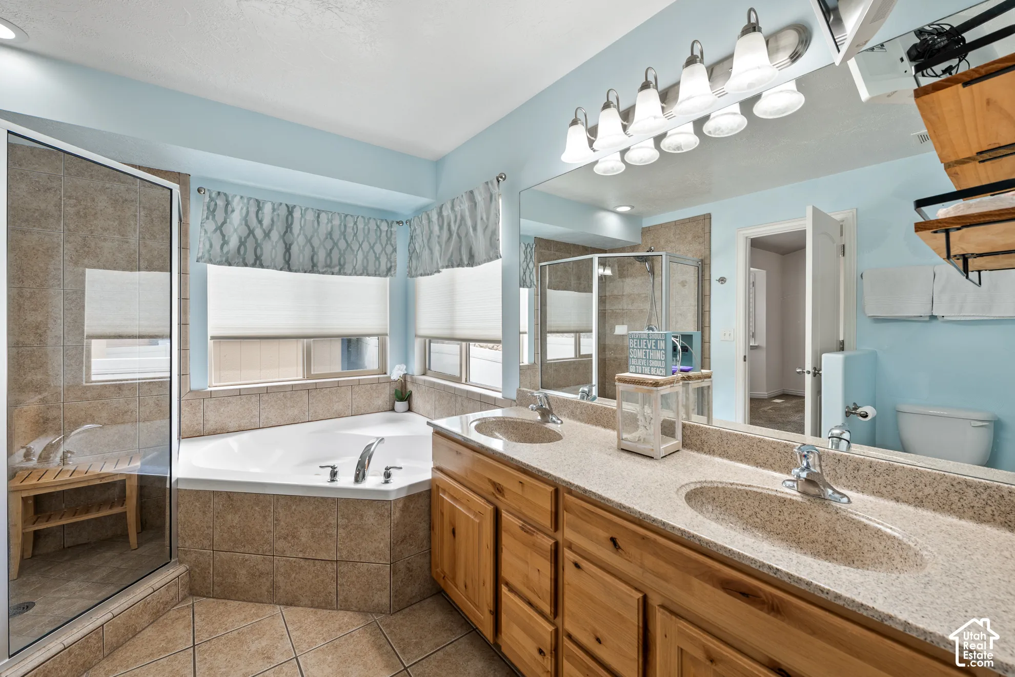 Dual sink vanity with wood-finish cabinetry and light-toned countertop, large wall-mounted mirror, corner soaking tub with tiled surround, and glass-enclosed shower with tiled walls