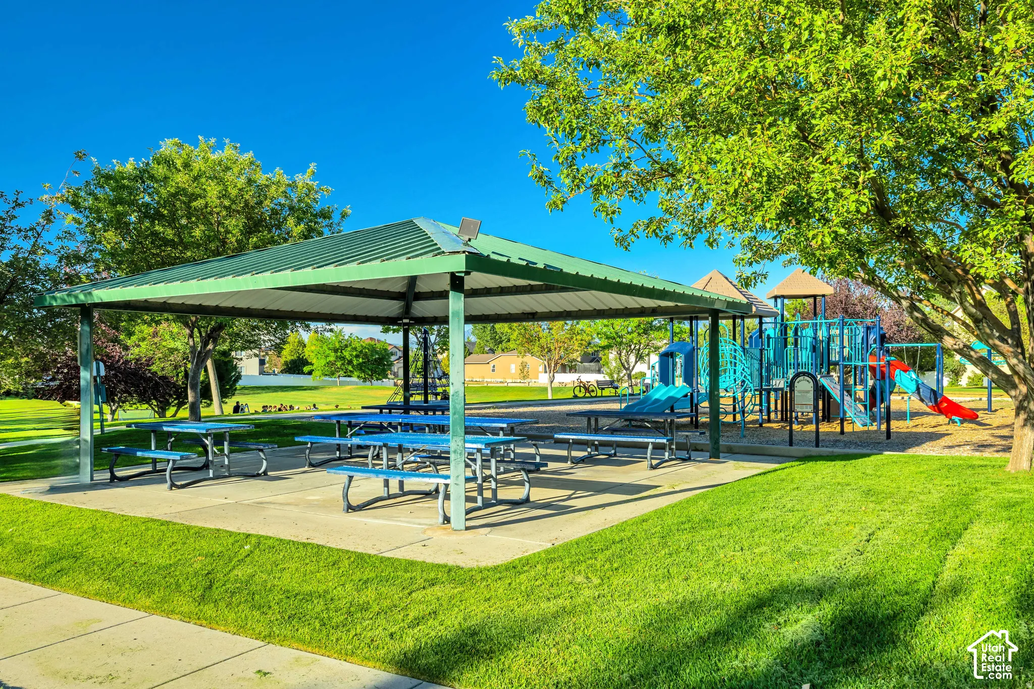 Expansive recreational area featuring a large covered picnic structure, multiple picnic tables, a vibrant playground, mature trees, and a manicured lawn