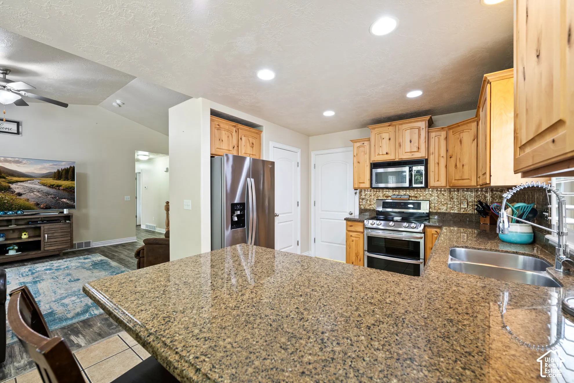 Kitchen featuring a double basin stainless steel sink, stainless steel appliances, wood-finish cabinetry, and a textured backsplash, with a stone-finish countertop extending into a breakfast bar