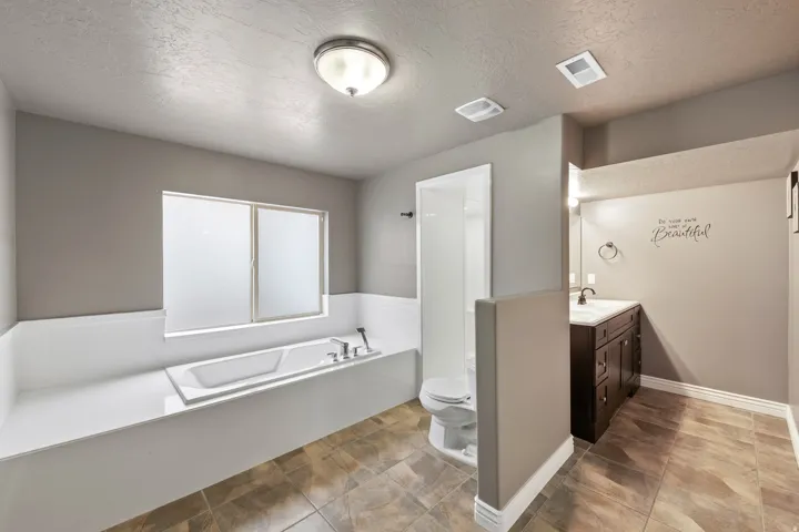 Soaking tub with a frosted window, separate shower enclosure, single vanity with dark wood cabinetry, and neutral-toned floor tiling