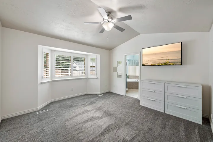 Vaulted ceiling with a ceiling fan, bay window with plantation shutters, and carpet flooring