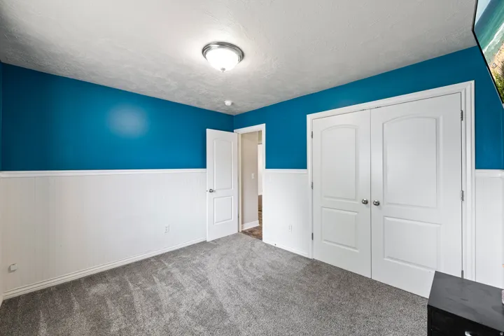 Plush gray carpeting, white wainscoting, vibrant blue wall paint, double-door closet, and an overhead flush-mount light fixture