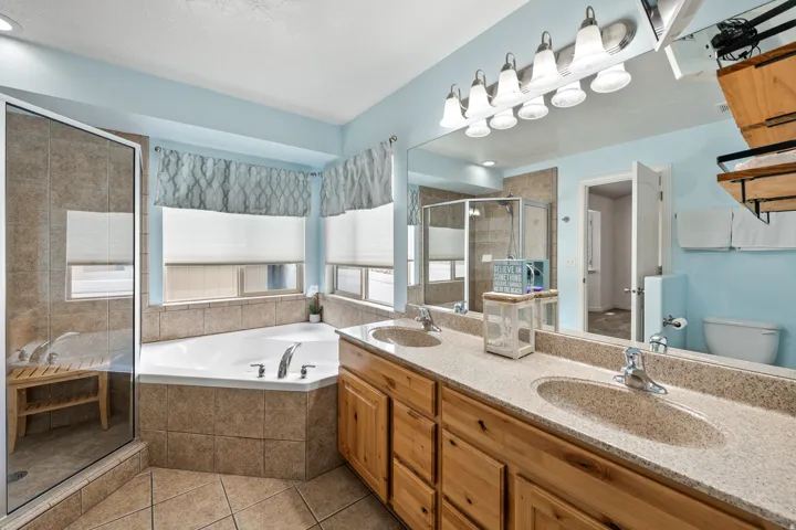 Dual sink vanity with wood-finish cabinetry and light-toned countertop, large wall-mounted mirror, corner soaking tub with tiled surround, and glass-enclosed shower with tiled walls