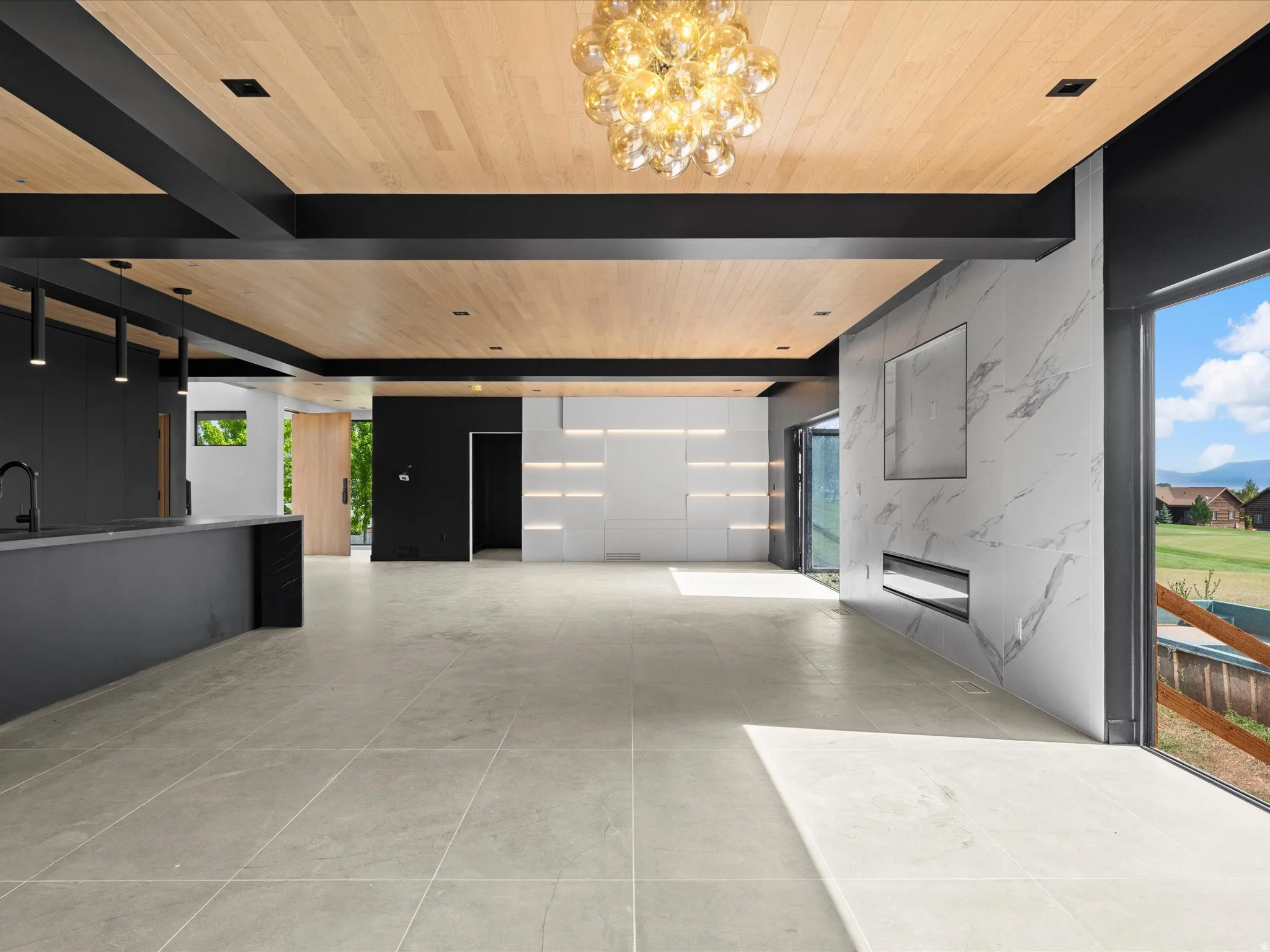Wood-paneled ceiling with exposed black beams, large format floor tiling, built-in linear fireplace, integrated wall shelving with accent lighting, and a large sliding glass door