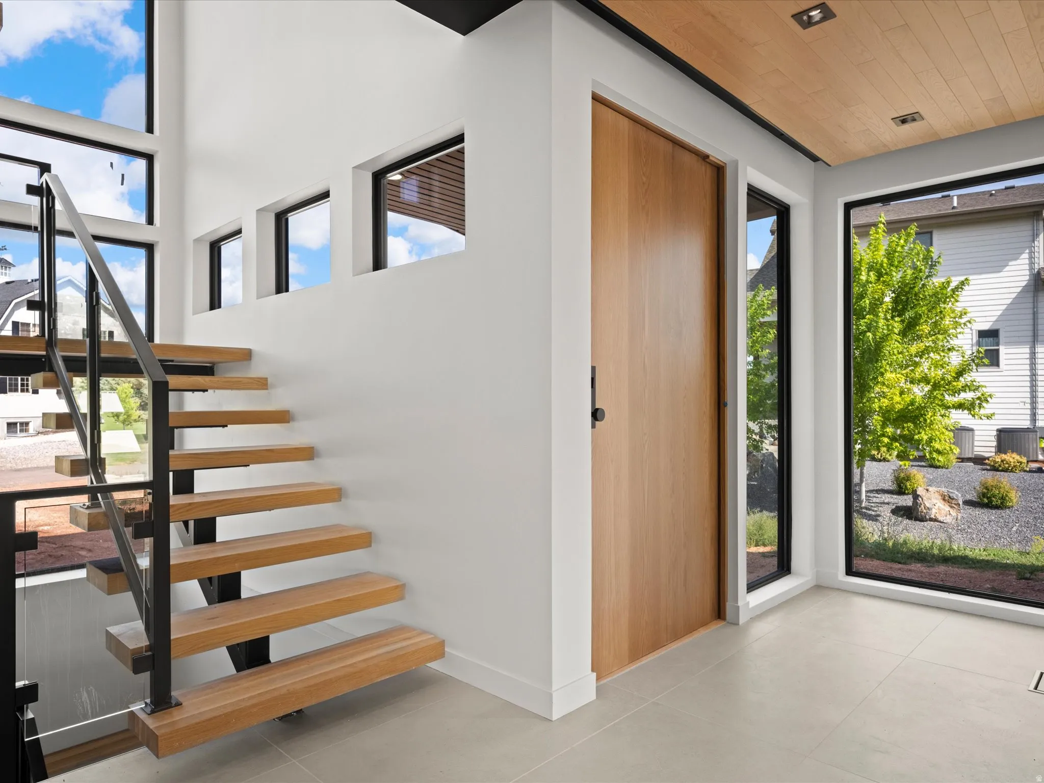 Floating wood staircase with metal and glass railing, natural wood-finish door, wood-paneled ceiling with recessed lighting, and large format floor tiling