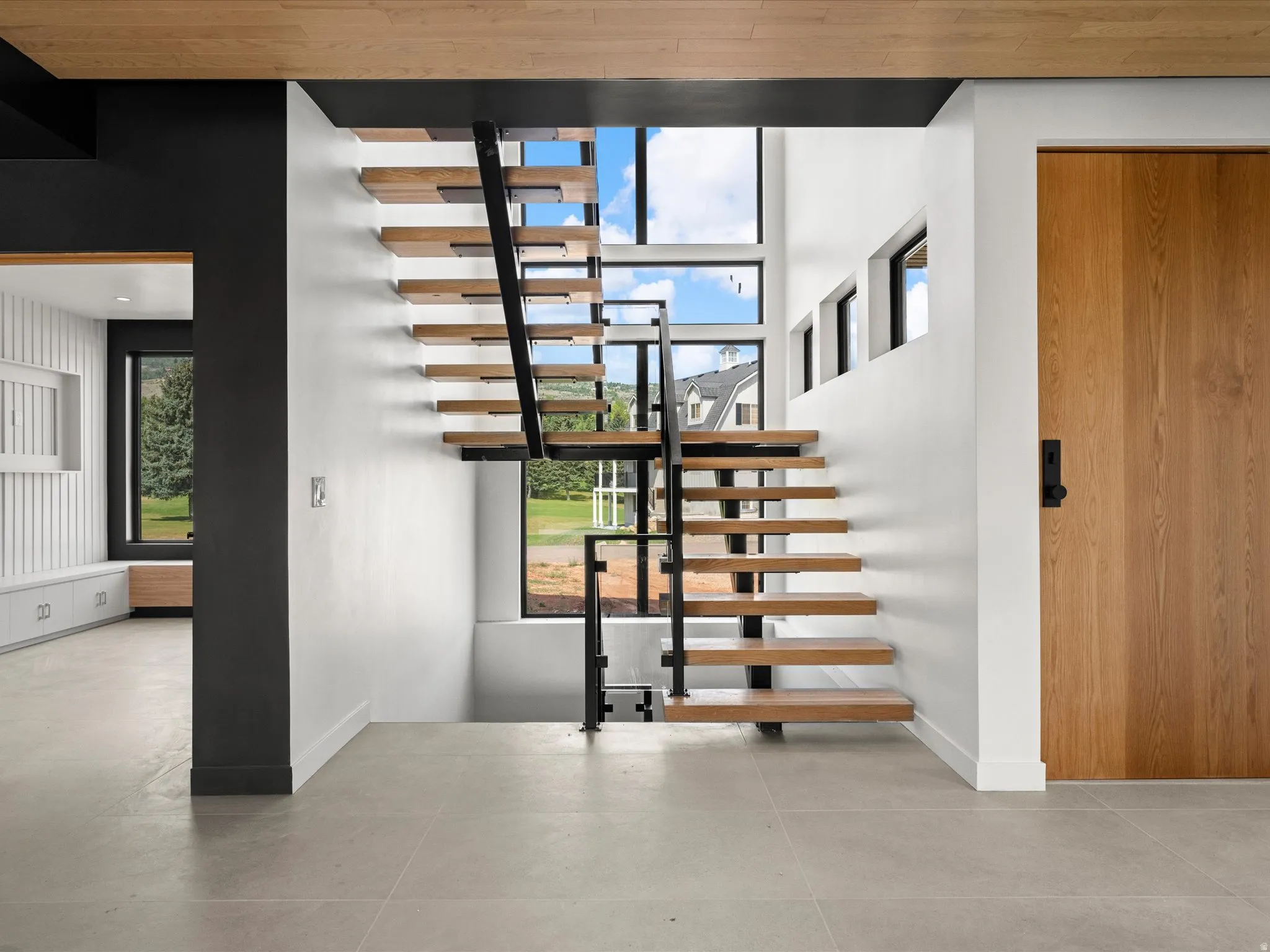 Cantilevered wood-finish staircase with black metal railing, large format tile flooring, and a natural wood-finish door