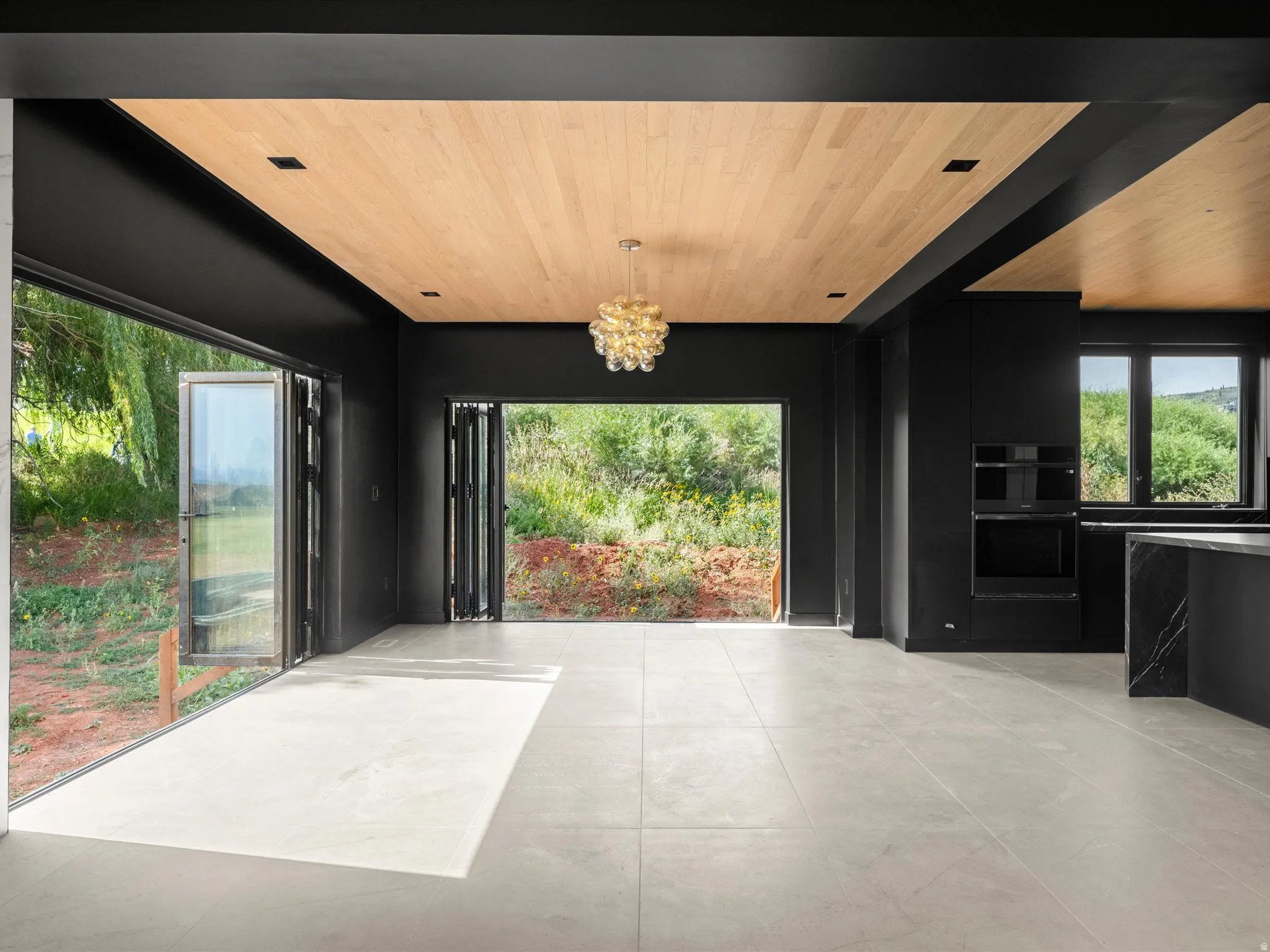 Wood-panel ceiling with recessed lighting, large format Arizona tile flooring, bi-fold glass doors, built-in wall oven, and a kitchen island with a stone slab waterfall countertop