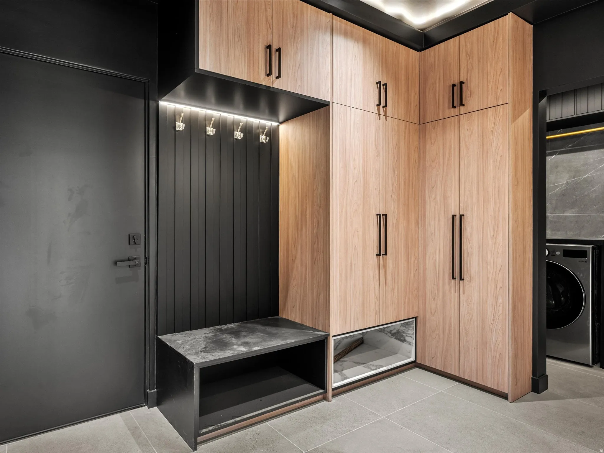 Wood-finish cabinetry with black hardware, integrated task lighting, black slat wall paneling, and a stone-finish bench