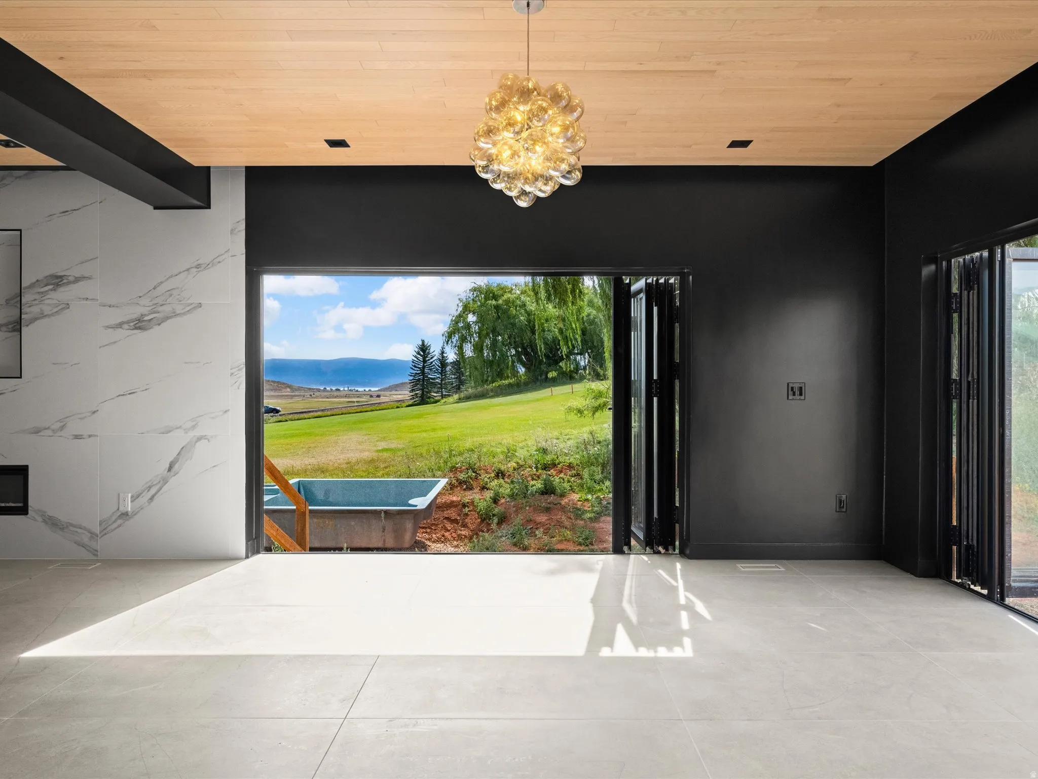 Light wood-finish ceiling, modern chandelier, recessed lighting, expansive folding glass doors, and light-toned tile flooring