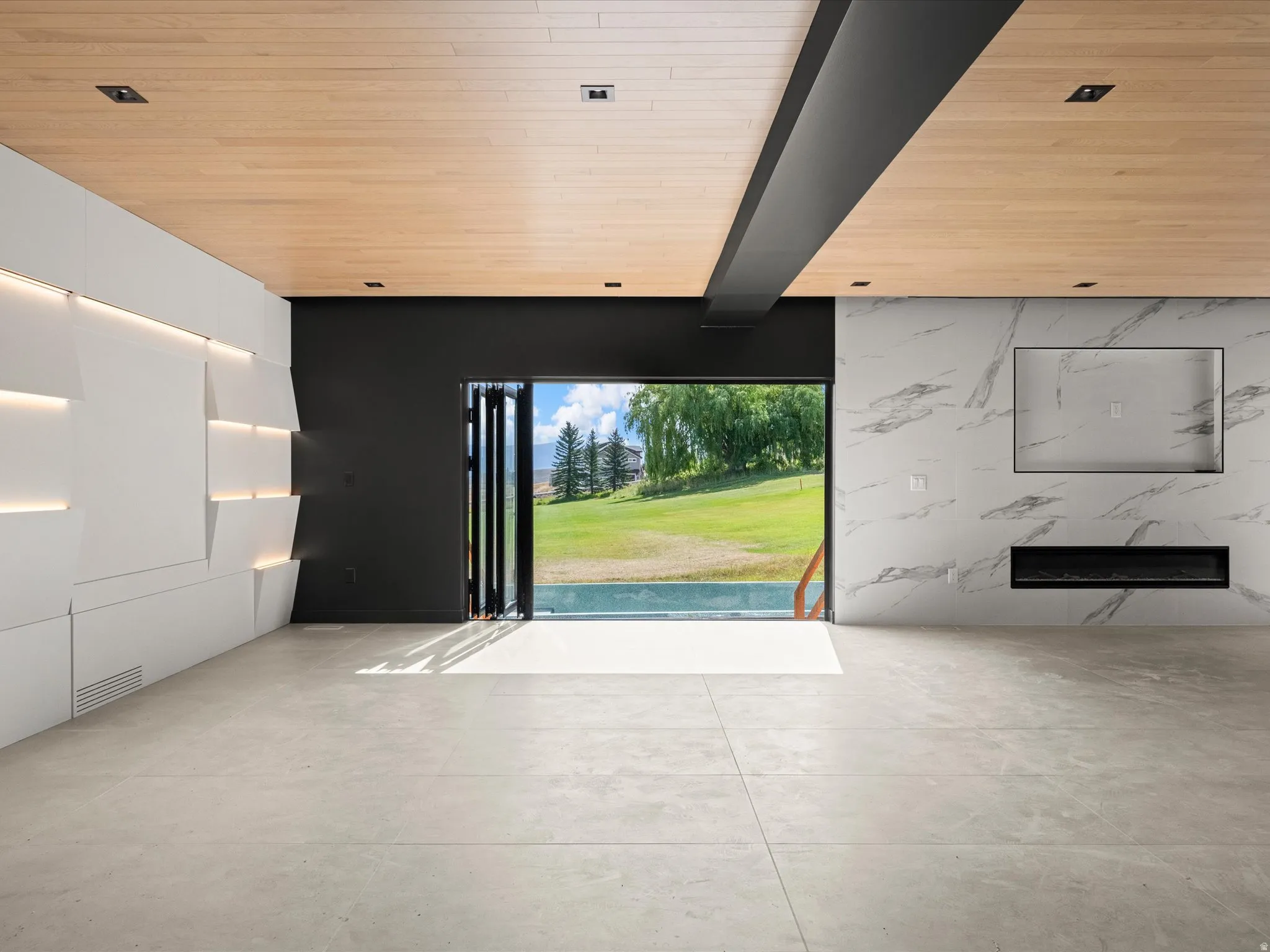 Expansive floor-to-ceiling bi-fold doors, wood-finish ceiling, white textured accent wall with integrated lighting, large format tile flooring, and a stone-finish accent wall with a built-in fireplace