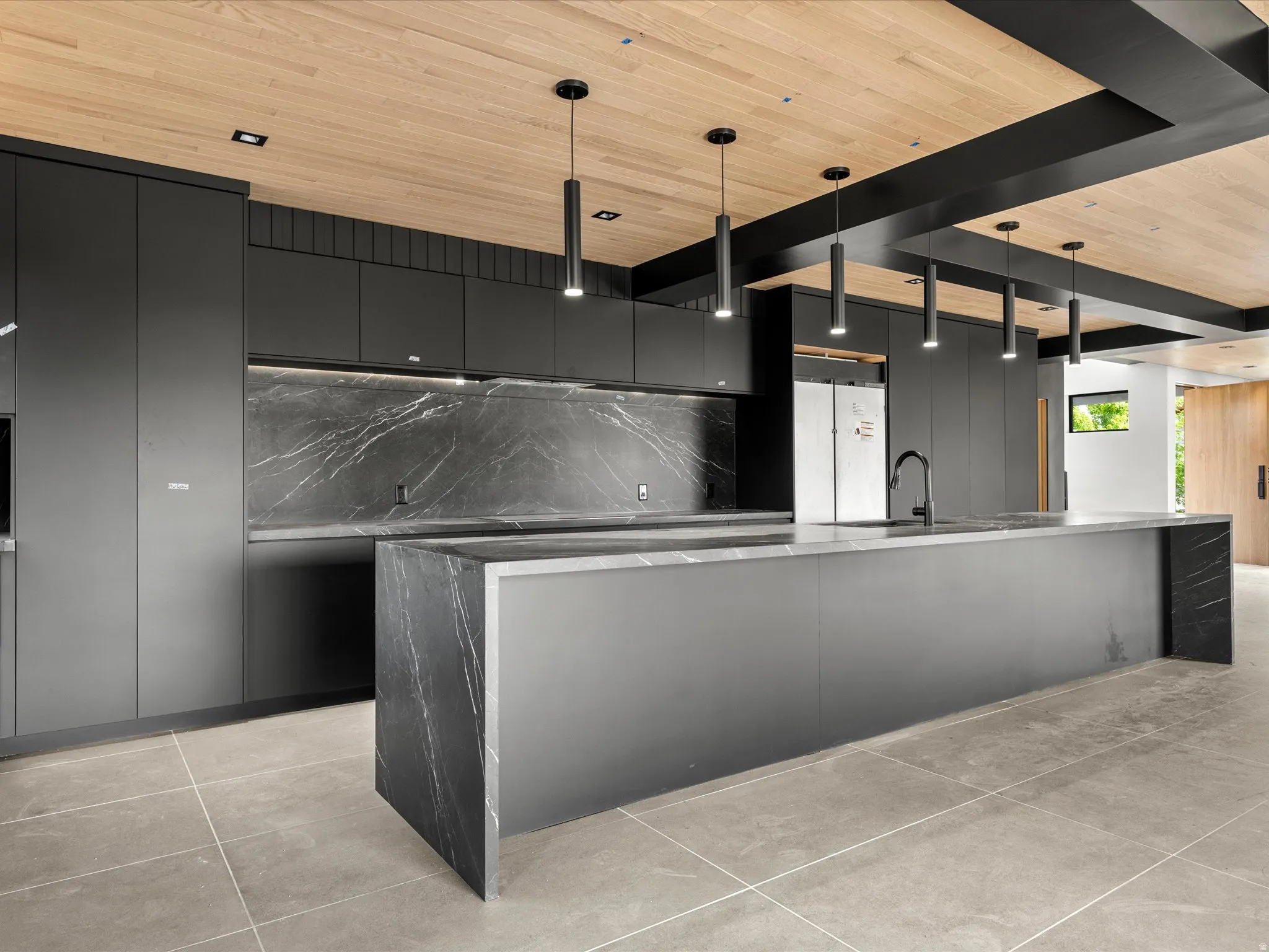 Sleek kitchen featuring dark-toned cabinetry, a waterfall island, elegant marble-patterned slab backsplash, black pendant lighting, and a wood-paneled ceiling