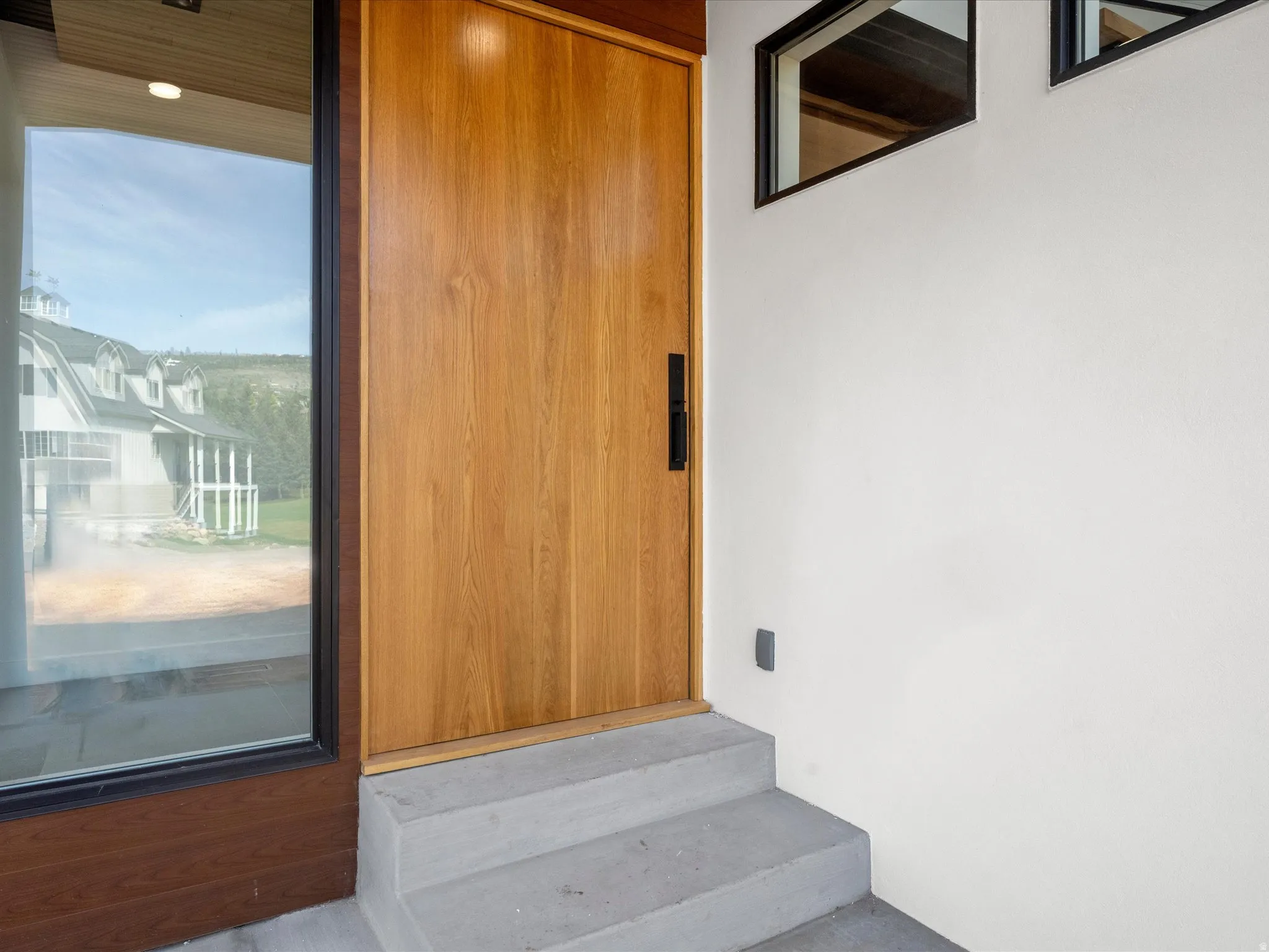 Wood-finish entryway pivot door with a black handle, flanked by a full-height window and transom windows