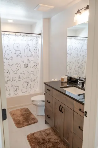 White tiled flooring, light wood-finish vanity with dark hardware, dark countertop, single-basin undermount sink, and a mirrored wall