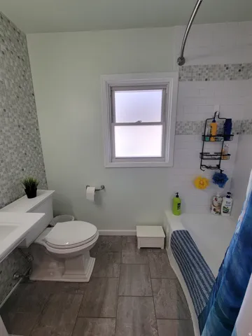Bathroom featuring gray-toned tile flooring, mosaic tile accent wall, white window frame, and a built-in bathtub with subway tile surround