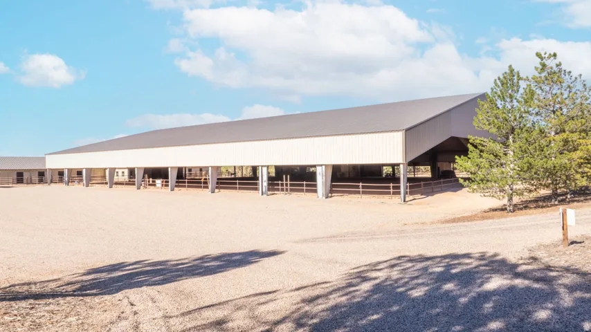 Expansive covered equestrian arena with metal siding and roof, featuring an attached barn structure and an outdoor training area