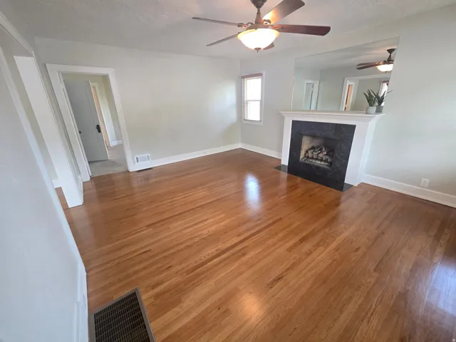 Hardwood flooring, fireplace with dark stone surround, white mantel, ceiling fan with integrated lighting, and a mirrored wall accent