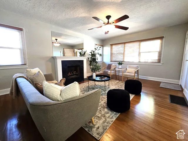 Living area featuring wood-finish flooring, fireplace with a dark stone surround, oversized wall mirror, ceiling fan with integrated lighting, and large windows