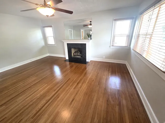 Warm wood flooring, ceiling fan with integrated lighting, fireplace with dark tile surround, white mantel, and multiple windows