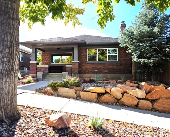 Brick exterior featuring a covered front porch, stone retaining wall, concrete walkway, and masonry chimney