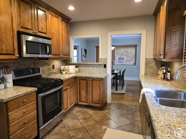 Kitchen featuring warm-tone wooden cabinetry, stone countertops, stainless steel appliances, dual-basin sink, and tile flooring