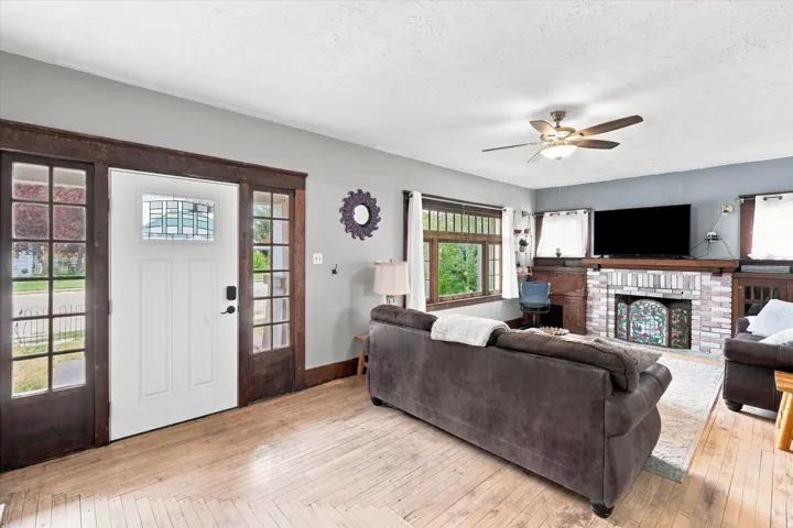 Wood-finish flooring, white entry door with stained glass transom, dark wood-trim sidelights, stone fireplace surround, and ceiling fan