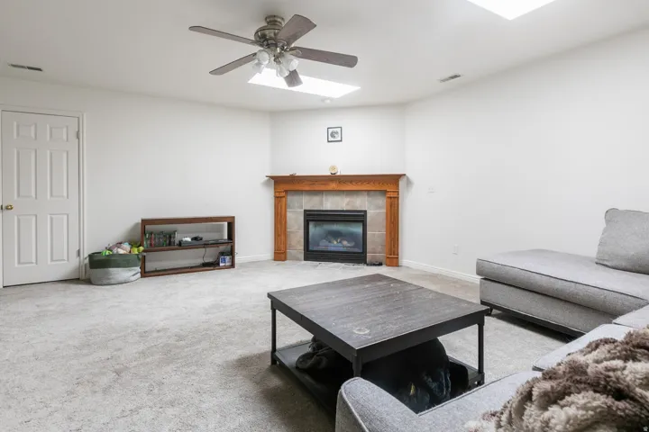 Carpeted living area featuring a corner fireplace with a wood mantle and tile surround, two skylights, and a ceiling fan