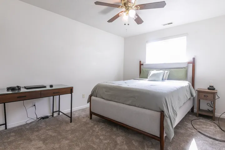 Plush carpet flooring, ceiling fan with integrated lighting, and a large window with blinds
