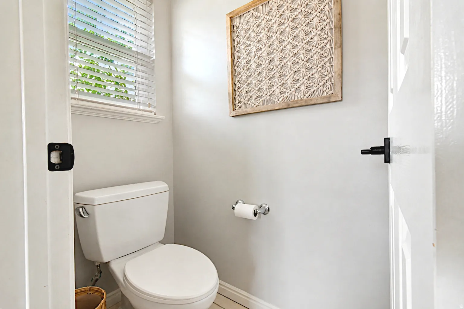 White toilet with chrome flush lever, window with blinds, light-toned wall paint, and matte black door hardware
