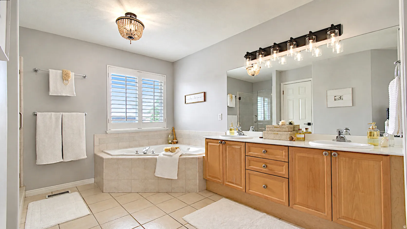 Tiled corner soaking tub with window shutters, dual vanity with light wood cabinetry, white countertops, and expansive wall mirror with multi-bulb light fixture