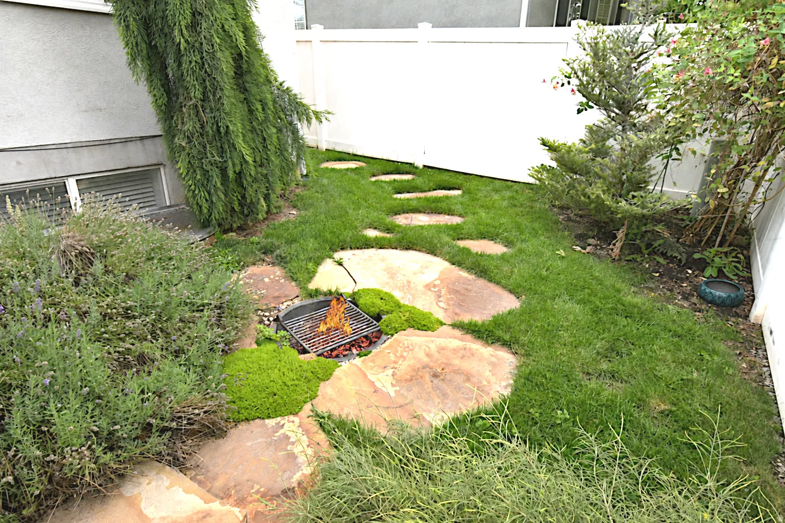 Landscaped side yard featuring a fire pit, stepping stone path, mature greenery, and a white privacy fence