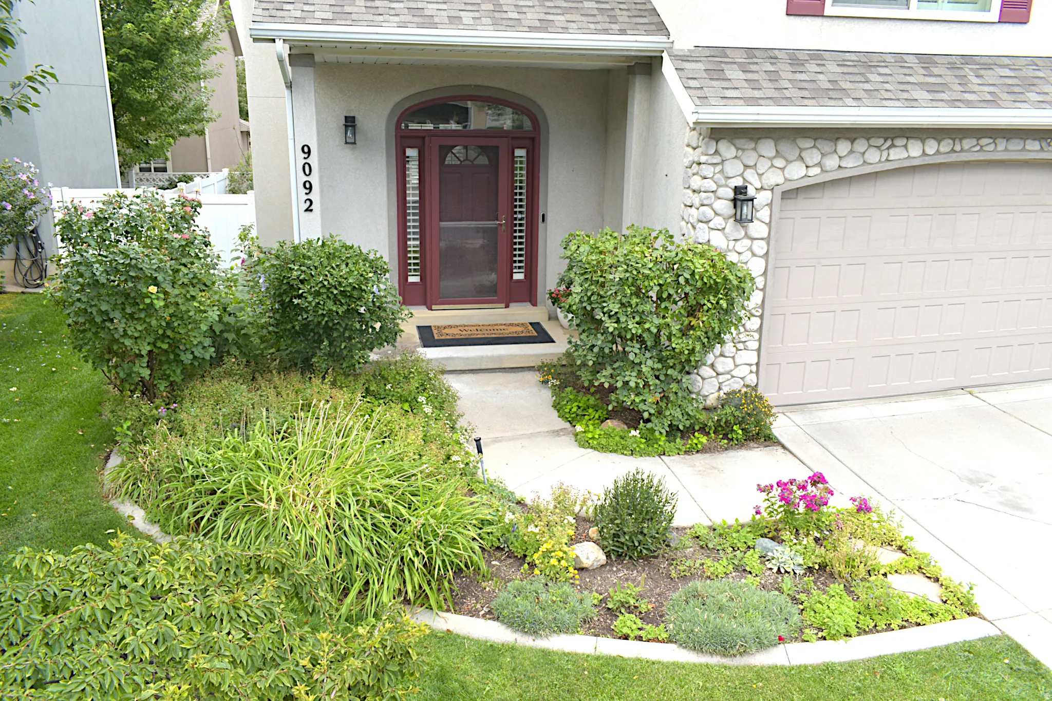 Arched entry with a red door, stone veneer garage wall, concrete driveway, and established landscaping